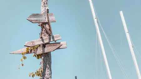 Verwitterter Wegweiser mit leeren Schildern vor Segelmasten und blauem Himmel